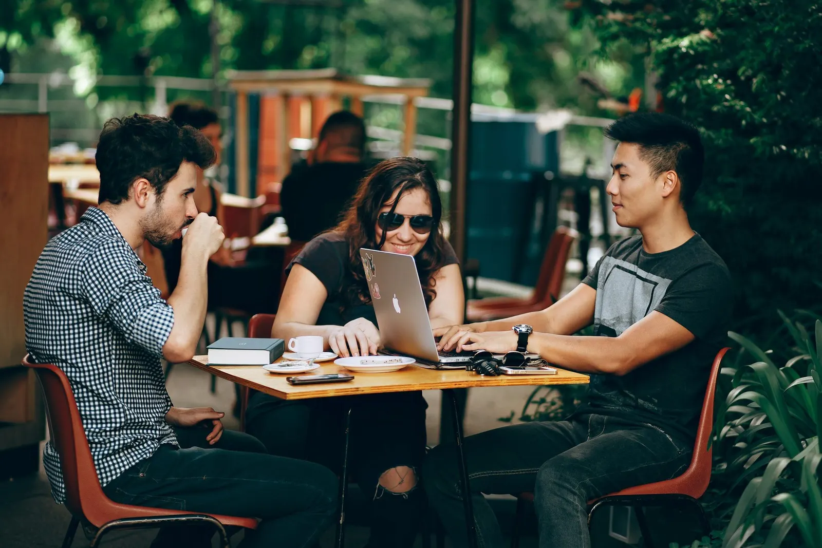 Three friends at a sunlit café table, a laptop open between them, mid-conversation.