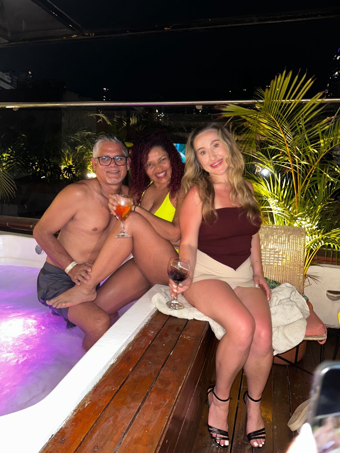 Three guests smiling by the rooftop jacuzzi at night with drinks.