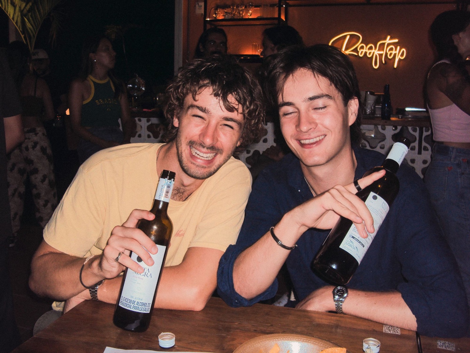 Two friends laughing over bottles of wine at the rooftop bar, the neon Rooftop sign behind.