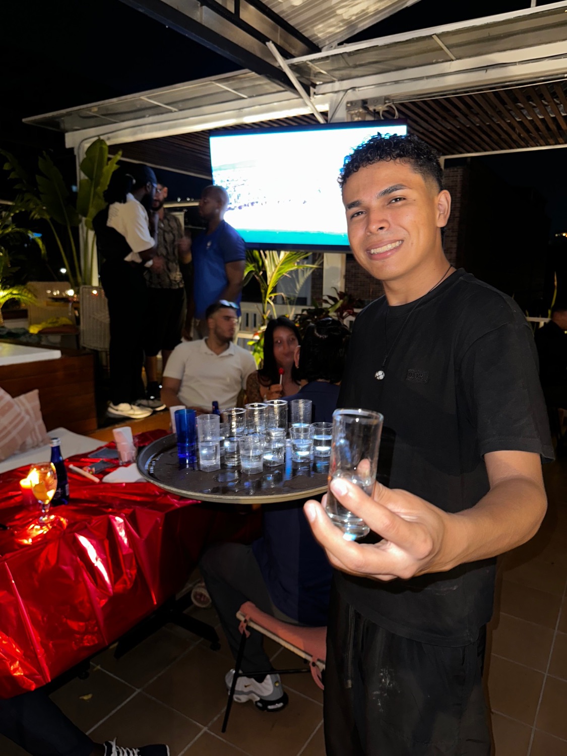 A smiling host offering a shot from a full tray on the rooftop.