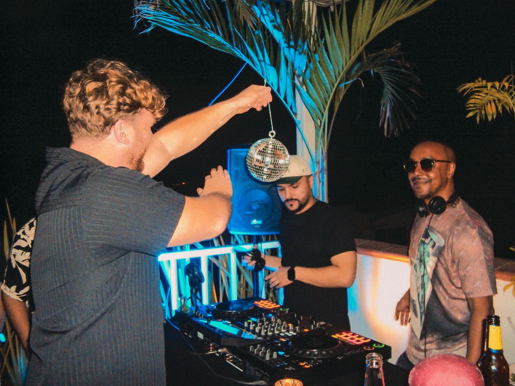 A DJ on the rooftop at night, a mirrorball held up beside the decks.