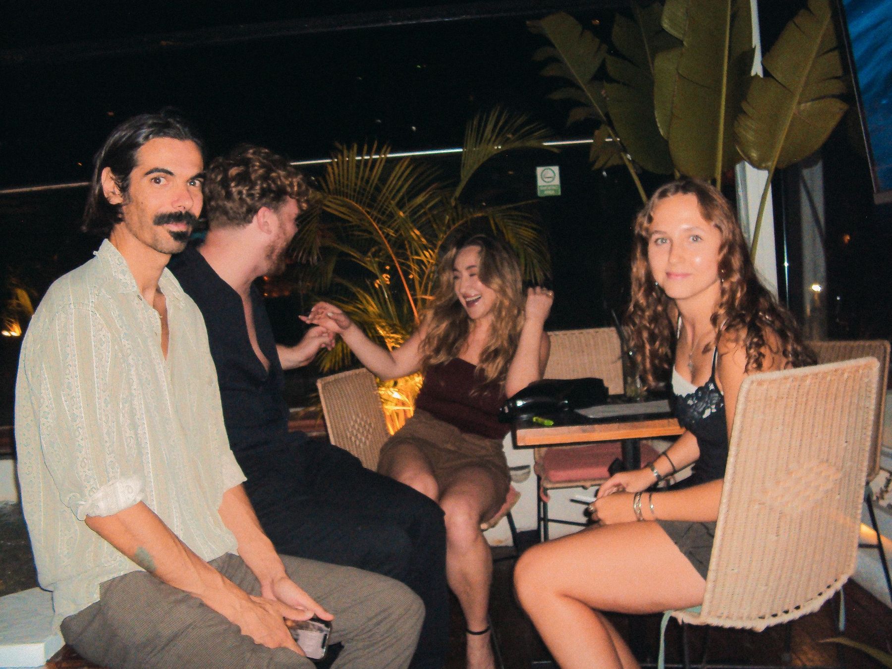 The owner laughing with guests around a table on the rooftop at night, palms lit behind them.