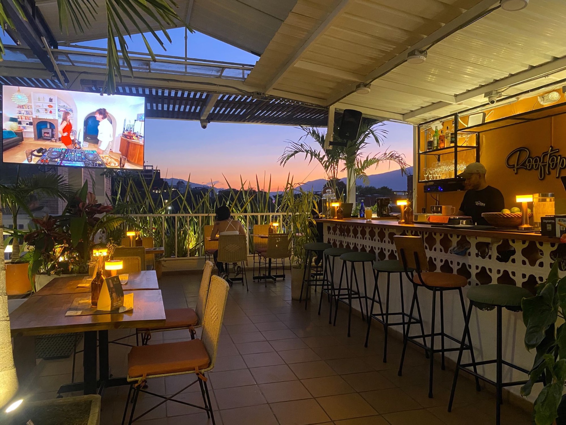 The rooftop bar at sunset, lamps lit on the tables and the hills behind.