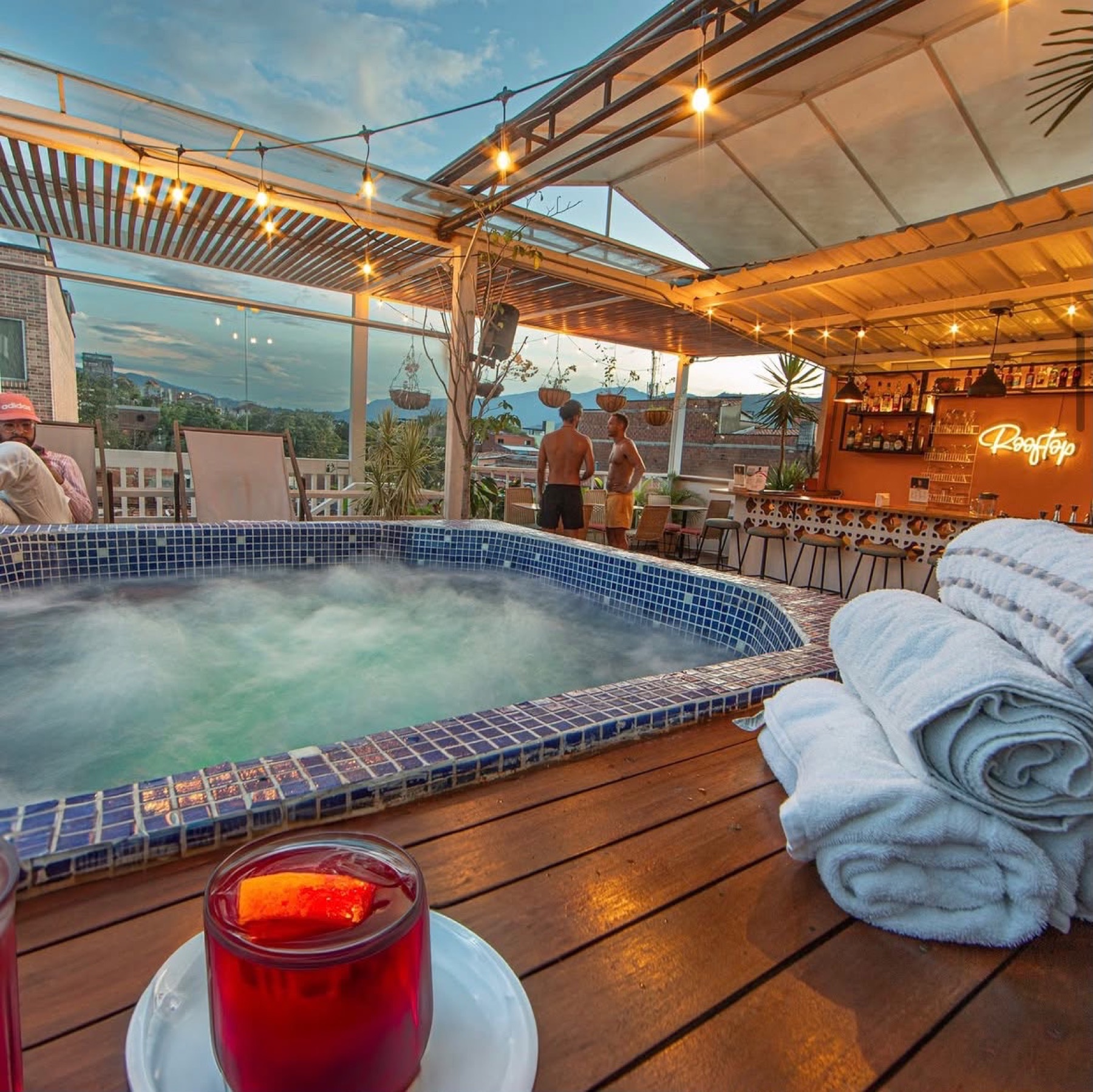 The tiled rooftop jacuzzi steaming at sunset, towels and a drink on the deck, the neon Rooftop bar behind.