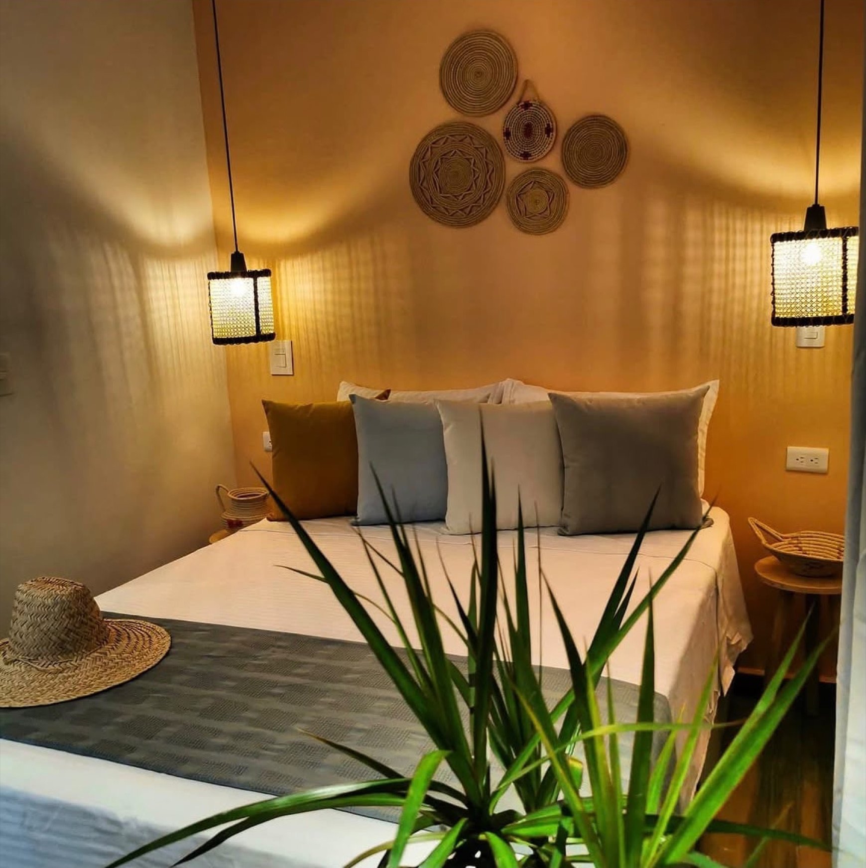 A made bed under woven wall baskets, rattan pendant lamps lit either side.